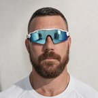 A close-up portrait of a man with a beard wearing white wraparound sunglasses with blue mirrored lenses, set against a plain light gray background. TEROVIK APEX sport sunglasses