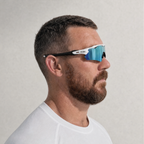 A man in profile wearing TEROVIK APEX wraparound sunglasses with a white frame, black temples, and blue mirrored lenses, set against a neutral light gray background.