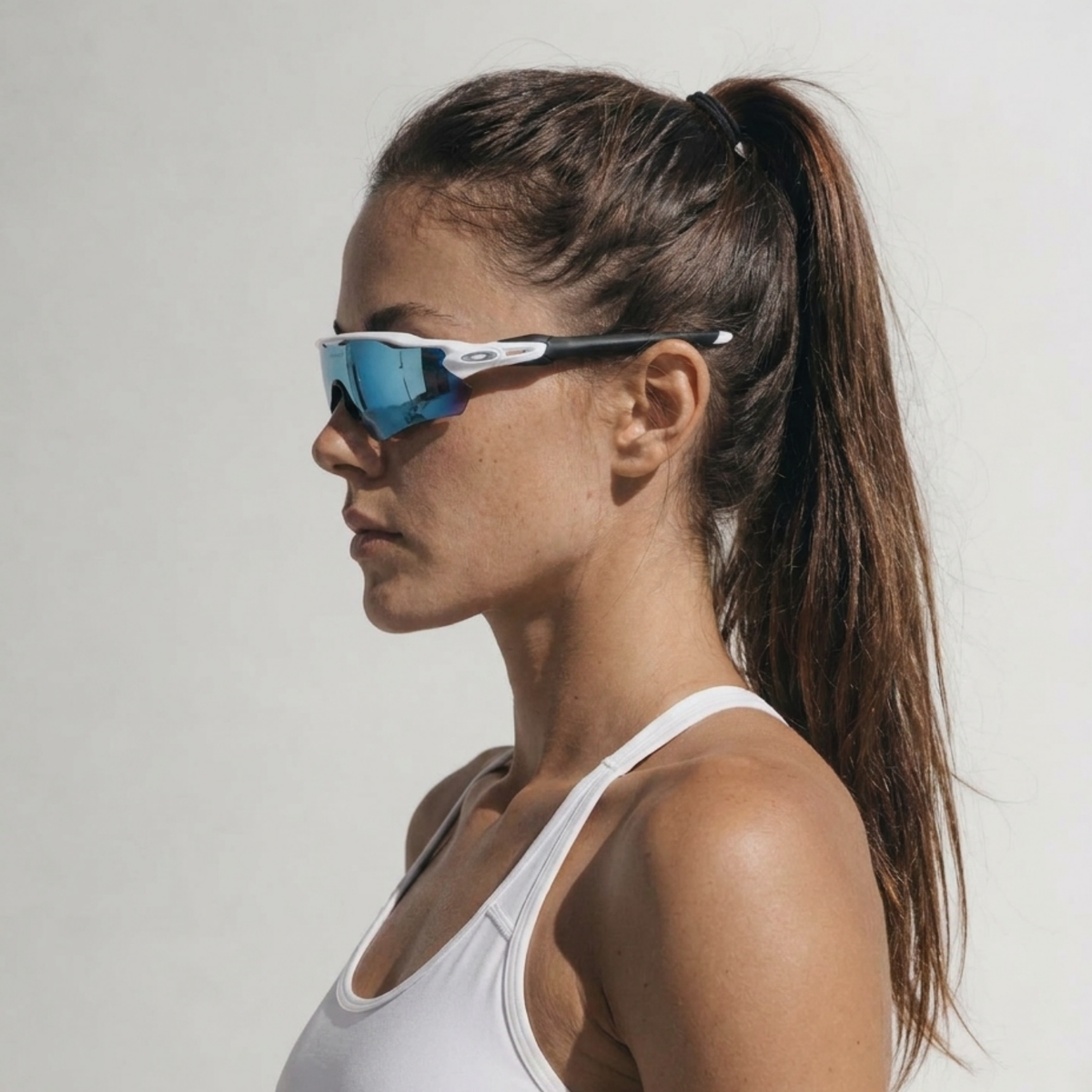 A woman in profile wearing TEROVIK APEX sport sunglasses with a white frame and blue mirrored lenses, paired with a white sports bra against a plain light gray background.
